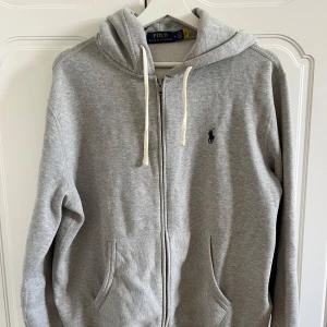 Grå hoodie från Ralph Lauren - Säljer en stilren grå hoodie från Ralph Lauren med dragkedja och justerbar huva. Den har en broderad logga på bröstet och är tillverkad i mjukt material för extra komfort. Perfekt för en avslappnad look.