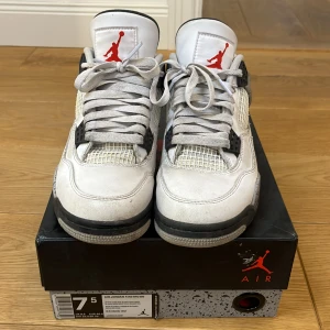 Nike Air Jordan 4 Retro white cement  - Säljer ett par klassiska Nike Air Jordan 4 Retro sneakers i vit och grå färg med svarta detaljer. Skorna har snörning och den ikoniska Jumpman-loggan i rött på plösen. Perfekta för sneakerheads som älskar retrostil.