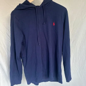 Mörkblå hoodie från Ralph Lauren - Säljer en stilren mörkblå hoodie från Ralph Lauren med det klassiska röda logotypbroderiet på bröstet. Tröjan har långa ärmar och en bekväm huva med dragsko. Perfekt för en avslappnad look.