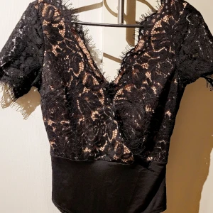Svart spetsblus med kort ärm - Elegant svart spetsblus (body) med korta ärmar och v-ringning. Blusen har en omlott-design och är dekorerad med spetsdetaljer som ger en sofistikerad look. Perfekt för en kväll ute eller en speciell tillställning.
