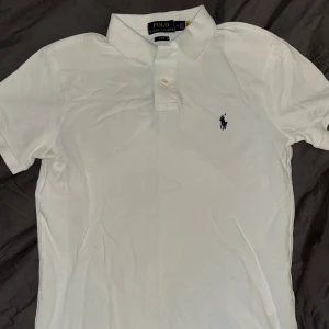 Vit pikétröja från Ralph Lauren - Säljer en klassisk vit pikétröja från Ralph Lauren i slim fit. Tröjan har korta ärmar och en broderad logga i blått på bröstet. Perfekt för en stilren look. Nästan oanvänd. Om man har frågor är det öppet att ställa dem.