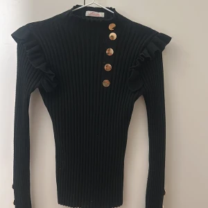 Svart ribbad tröja med knappar - Snygg svart ribbad tröja från Raglady med guldknappar längs axeln och ärmarna. Tröjan har volangdetaljer vid axlarna och långa ärmar. Perfekt för en stilren look.