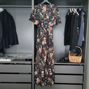 Blommig lång jumpsuit med fågelmotiv - Säljer en elegant jumpsuit med ett vackert blommigt mönster och fågelmotiv. Klänningen har korta ärmar och en omlott-design som ger en fin passform. Det jag gillar med denna är att den passar året runt!🌸
