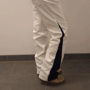 Vita byxor med mörkblå detaljer - Unika vita byxor med mörkblå sidopaneler och en stor mörkblå rosett baktill. Byxorna har en straight passform och är tillverkade i ett jeansmaterial. Perfekta för dig som vill sticka ut med en cool och annorlunda stil.