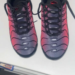 Nike Air Max Tn i röd, blå och dvart - Snygga Nike Air Max Tn sneakers i en cool röd och svart färgkombination. Skorna har en unik design med svarta vågformade detaljer och synlig Air Max-dämpning i sulan. Perfekta för en sportig look.