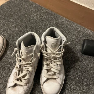 Vita Nike sneakers - Säljer ett par vita Nike sneakers med högt skaft och klassisk snörning. Skorna har en stilren design med Nike-loggan på sidan. Perfekta för en sportig look.
