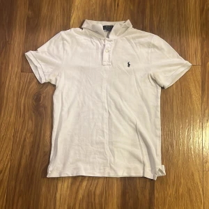 Vit kortärmad pikétröja från Ralph Lauren - Säljer en klassisk vit pikétröja från Ralph Lauren med korta ärmar och en broderad logga på bröstet. Tröjan har en stilren design med knappar vid halsen och är perfekt för en avslappnad look.