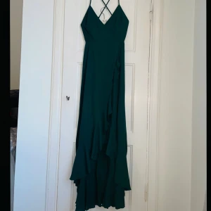 långklänning - Elegant grön långklänning med smala axelband och volangdetaljer. En perfekt klänning för lite finare tillställningar med sin flytande design och djupa färg. Klänningen har en v-ringad fram och korsade band i ryggen. Är en storlek L men skulle säga att den passar xs-