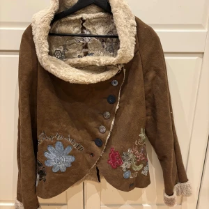 Brun mockajacka från Desigual - Unik brun mockajacka från Desigual med broderade blommor och mönster. Jackan har en mysig fuskpälsfoder och knappar framtill. Perfekt för att sticka ut med sin kreativa design och detaljer.