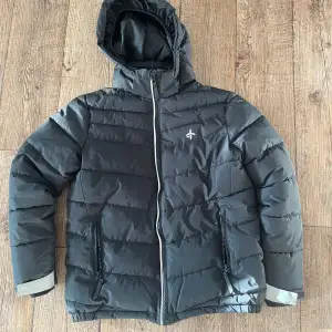 Säljer en svart pufferjacka från Cross Sportswear med huva och dragkedja. Jackan har två praktiska fickor med dragkedjor och är perfekt för kyligare dagar. 