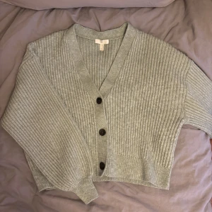 Grön ribbad kofta från H&M - Säljer en stilren grön ribbad kofta från H&M med knappar framtill. Koftan har en v-ringad design och långa ärmar, perfekt för lager-på-lager under kyligare dagar. Passar både till jeans och kjol för en avslappnad look.