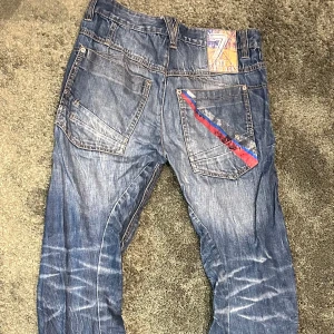 Blå jeans med unika detaljer - Snygga blå jeans med coola detaljer från Kappahl. De har en unik design med en röd och blå rand på bakfickan och ett mönstrat innerband i midjan. Dragkedja på ena fickan ger en extra edge. Perfekta för dig som vill sticka ut!
