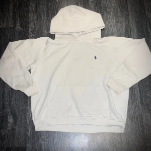 Vit hoodie från Polo Ralph Lauren - Säljer en klassisk vit hoodie från Polo Ralph Lauren med en liten blå logga på bröstet. Den har en stor ficka framtill och en bekväm huva. Perfekt för en avslappnad stil.