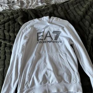 Emporio armani - Säljer en stilren vit hoodie från Emporio Armani med EA7-logga på framsidan. Tröjan har långa ärmar och en praktisk huva. Perfekt för en avslappnad look.
