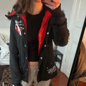 Svart dunjacka från Ed Hardy - Säljer en svart dunjacka från Ed Hardy med röd insida och broderade detaljer. Jackan har en stor huva med fuskpäls och tryck på ryggen och ärmen. en del av dragkedjan är lite lös men går att sy ihop lätt, detta gör så att det är lite svårare att stänga. Storlek M på lappen men skulle säga att den sitter som S. Pris är diskuterbart