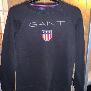Svart tröja från GANT - Säljer en svart tröja från GANT med broderad logotyp och emblem på framsidan. Tröjan har långa ärmar och rund halsringning. Perfekt för en stilren och bekväm look. Barnstorlek 