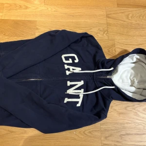 Mörkblå hoodie från GANT - Säljer en snygg mörkblå hoodie från GANT med dragkedja och vit text på framsidan. Den har en justerbar huva med vita snören och en mjuk insida för extra komfort. Perfekt för en avslappnad stil.