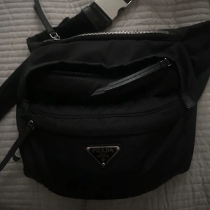 Svart axelväska från Prada - Snygg svart axelväska från Prada med en stilren design. Väskan har en praktisk dragkedja och det ikoniska Prada-emblemet på framsidan. Går att bära på ryggen och fram. Finns även så du kan justera bandet, vilken längd du vill ha.
