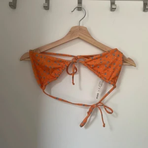Orange blommig bikiniöverdel - Snygg orange bikiniöverdel med blommigt mönster och knytband i nacken och ryggen. Perfekt för soliga dagar på stranden eller vid poolen. Den har en klassisk trekantsform som ger en fin passform.