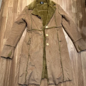 Beige mockakappa med foder - Snygg beige mockakappa med mjukt foder och knappar framtill. Kappan har långa ärmar och en klassisk krage. Perfekt för kyligare dagar och ger en stilren look. Oanvänd från B2 i storlek S! 