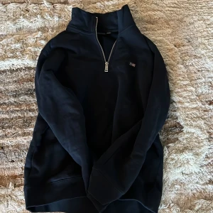 Svart tröja med dragkedja från Lexington - Säljer en stilren svart tröja från Lexington med halv dragkedja. Perfekt för kyligare dagar med sin bekväma passform och klassiska design. Tröjan har långa ärmar och en liten logga på bröstet. Passar ungdomar i åldern 12-14 år.