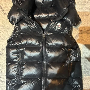 Svart dunväst från Moncler - Snygg svart dunväst från Moncler med dragkedja och ikonisk logga på bröstet. Perfekt för kyligare dagar med sin quiltade design och stilrena look. Västen har en praktisk huva och två fickor med dragkedja.