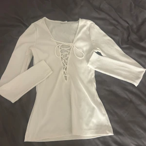 Vit ribbad topp från Gina Tricot - Säljer en stilren vit ribbad topp från Gina Tricot. Toppen har långa ärmar och en snygg snörning framtill som ger en v-ringad look. Perfekt för att skapa en chic och avslappnad stil.