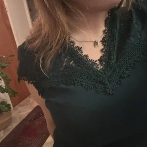 Mörkgrön topp med spetsdetaljer - Säljer en elegant mörkgrön topp med vacker spets runt halsringningen. Perfekt för både vardag och festliga tillfällen. Toppen har en normal passform. Ett måste i garderoben för den som älskar stilrena plagg! 