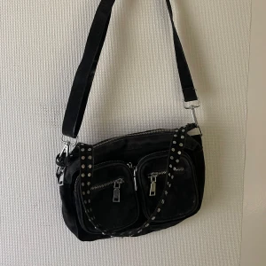 Svart axelväska från Noella - Svart axelväska från Noella med nitar på axelremmen🖤 Den har flera dragkedjefickor vilket gör det lättare att hålla den organiserad🙏 Bra men använt skick och har en längre justerbar rem som går att ta av och på