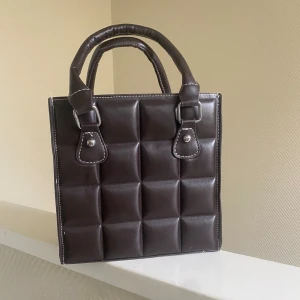 Brun quiltad handväska - Säljer en superfin brun handväska i quiltad stil. Den har ett coolt rutmönster och är gjord av syntetmaterial. Aldrig använd. Väskan har två handtag och en praktisk dragkedja upptill. Axelband kan tas på och av som man önskar. Perfekt för både vardag och fest!