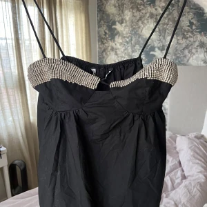 Svart klänning med pärldetaljer - Snygg svart topp med tunna axelband och pärldetaljer vid bysten. Perfekt för festliga tillfällen! Klänningen har en elegant och stilren design som passar bra till både nyår och andra speciella tillfällen.