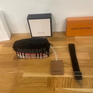 Svart axelväska från Burberry - Säljer en helt ny snygg svart axelväska från Burberry med klassiskt rutigt mönster i rött, vitt och grått. Väskan har en praktisk dragkedja och justerbar rem. Perfekt för både vardag och fest! 🖤