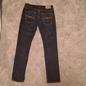 Mörkblå jeans från Nudie Jeans - Säljer ett par snygga mörkblå jeans från Nudie Jeans i bra skick. De har en klassisk femficksdesign med orange sömmar och en snygg logga på bakfickan. Perfekta för både vardag och fest! Passar till allt och är superbekväma. De är som nya och använda 2 gånger de är i storleken W 31, L32