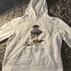Vit hoodie från Ralph Lauren med Polo Bear - Säljer en superfin vit hoodie från Ralph Lauren med den ikoniska Polo Bear som åker skidor. Perfekt för vintern och riktigt mysig med sin mjuka insida. Den har en normal passform och är i mycket bra skick. Perfekt för både vardag och chill.