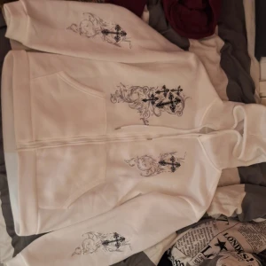 Vit hoodie med tryck från Romwe - Säljer en vit hoodie från Romwe i storlek M. Den har ett snyggt tryck med kors och blommor på framsidan. Hoodien har dragkedja och fickor, perfekt för en avslappnad stil. Materialet känns mjukt och skönt, perfekt för kyliga dagar. Står medium i storlek men känns som Large.