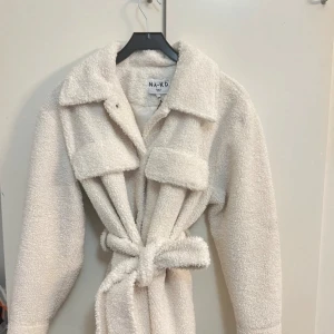 Beige teddyjacka från NA-KD - Supermysig beige teddyjacka från NA-KD i storlek M. Perfekt för höst och vinter med sitt varma material och snygga bälte i midjan. Jackan har långa ärmar och en klassisk krage. Den är i mycket bra skick och redo för nya äventyr!