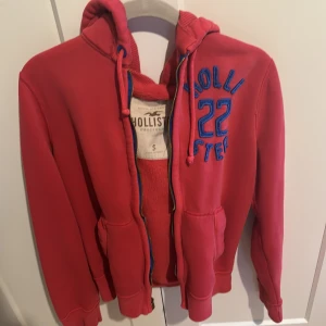 Röd hoodie från Hollister - Säljer en snygg röd hoodie från Hollister i storlek S. Den har en mysig fodrad insida och en stor luva. Perfekt för kyliga dagar! Den har dragkedja och fickor framtill samt blå text på bröstet.