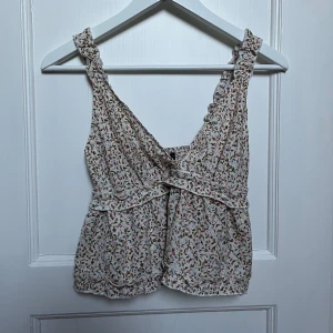 Blommig ärmlös topp - Söt blommig topp med volangdetaljer och smockad rygg. Perfekt för sommardagar! Den har en luftig passform och är i ett lätt material med små blommor i rött och grönt på en vit bakgrund.