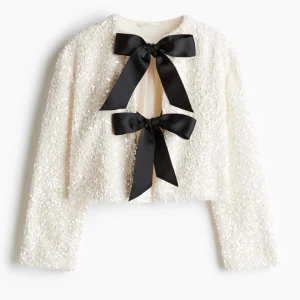 Viral vit paljettjacka med svarta rosetter från H&M stl S - Säljer en superfin vit paljettjacka med två stora svarta rosetter framtill. Jackan är croppad och har långa ärmar, perfekt för festliga tillfällen. Den glittriga ytan gör den till ett riktigt statement-plagg. Passar perfekt till nyår eller andra speciella tillfällen!