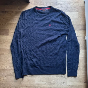 Mörkblå tröja från Morris - Säljer en snygg mörkblå tröja från Morris i 100% merinoull. Den har ett diskret kabelstickat mönster och en liten röd broderad logga på bröstet. Perfekt för både höst och vinter, med långa ärmar och ribbade muddar. Supermjuk och stilren!