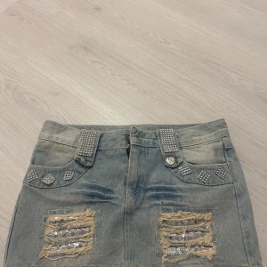 Jeanskjol med strassdetaljer - Snygg jeanskjol i storlek S, det står L men jag har storlek S och den passar mig. Coola strassdetaljer på fickorna och bältet. Kjolen har en sliten look med paljetter på de slitna delarna, vilket ger en trendig touch. Däremot så har dragkedjan gått sönder, men det går enkelt att fixa med ny dragkedja.🤗🤗