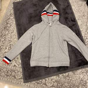 Säljer en snygg grå hoodie från Moncler i mycket bra skick. Den har en dragkedja framtill och coola ränder i rött, vitt och blått på huvan och ärmsluten. Perfekt för höst och vår när du vill vara både bekväm och stilren. Passar lika bra till vardags som till en chill festkväll.