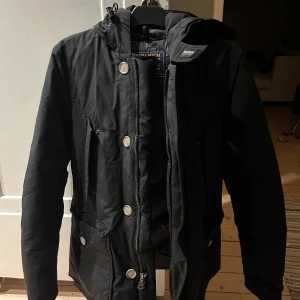 Svart Arctic Anorak från Woolrich - Säljer en svart jacka från Woolrich i bra skick. Den har en dragkedja och knappar framtill, perfekt för höst och vinter. Jackan är långärmad och har en stilren design med praktiska fickor. Perfekt för att hålla sig varm och snygg under kyliga dagar!