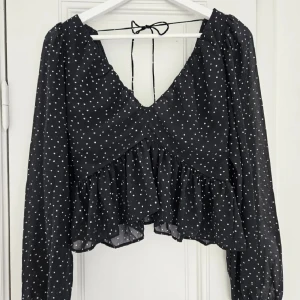 Svart blus med vita prickar - Säljer en supersöt svart blus med vita prickar. Den har långa ärmar och en volangdetalj nedtill. Blusen är v-ringad och har en knytning i ryggen. Perfekt för både vardag och fest!
