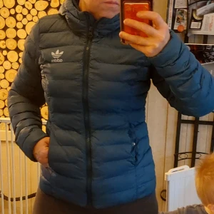 Blå dunjacka från Adidas - Säljer en snygg blå dunjacka från Adidas i storlek 34. Jackan har en figursyddl passform med dragkedja och huva, perfekt för kyliga dagar. Den är i mycket bra skick och har det klassiska Adidas-loggan på bröstet. Perfekt för höst och vinter!