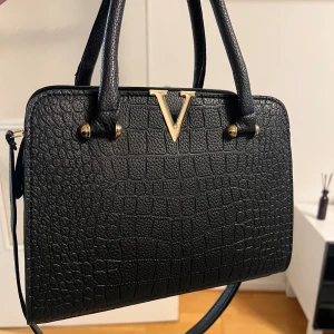 Svart handväska med krokodilmönster - Helt ny svart handväska i syntet med krokodilmönster och gulddetaljer. Väskan har en praktisk dragkedja och ett elegant V-emblem framtill.