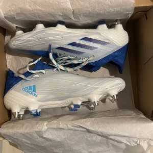Vita och blå fotbollsskor från Adidas - Säljer ett par snygga fotbollsskor från Adidas i vitt och blått. De är i nästan nyskick eftersom jag bara använt dem tre gånger och de har inga skador eller slitage. Jag säljer dem eftersom jag hellre vill ha fotbollsskor med strumpskaft. Skorna kommer med sex extra skruvdobbar, bra om du vill byta eller justera greppet.