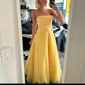 Gul axelbandslös balklänning - Säljer en fantastisk gul axelbandslös balklänning i mycket bra skick. Klänningen har en elegant drapering i midjan och är perfekt till bal. Den är köpt secondhand och har tidigare haft band som dock är avklippta. Jag hade den på min studentbal, se bilder. 