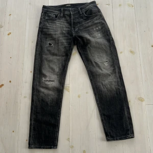 Svarta jeans från Jack & Jones - Säljer ett par svarta jeans från Jack & Jones i bra skick. De har en cool slitning på framsidan och en bekväm tapered passform. Perfekta för en avslappnad stil eller en utekväll. Midjan är normal och de är gjorda av slitstarkt denim.