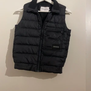 Svart dunväst från Calvin Klein Jeans - Säljer en snygg svart dunväst från Calvin Klein Jeans. Västen har en dragkedja framtill och en praktisk bröstficka med dragkedja. Perfekt för lager-på-lager under kyliga dagar. Passar både till vardags och lite mer uppklädda tillfällen.
